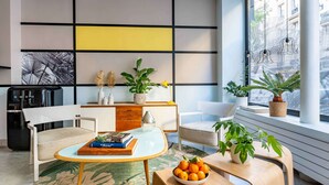 A modern living room with a sofa, a coffee table, and a dining table. There are potted plants and a bowl of fruit on the dining table. The room has a large window with a view of buildings outside. - Hôtel Palm - Astotel (Paris)