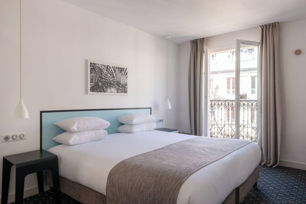 A hotel room with a bed, a nightstand, a wall-mounted light, a window with curtains, and a balcony view.