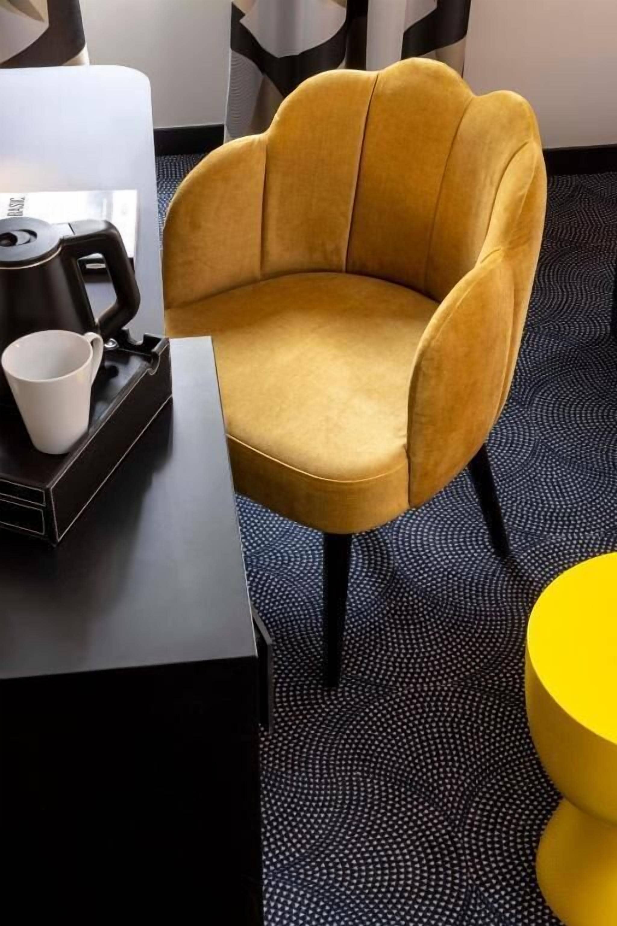 Una stanza con una poltrona giallo senape, un tavolino nero con una tazza bianca e un comodino giallo.
