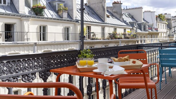 A balcony with a table set for breakfast, including croissants and orange juice, overlooking a row of Parisian buildings. - Hotel Régina Opéra Grands Boulevards (Paris)