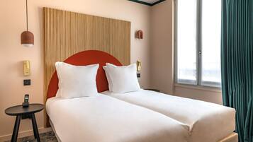 Une chambre d’hôtel avec deux lits, une petite table avec un téléphone, une lampe fixée au mur et une fenêtre avec des rideaux.