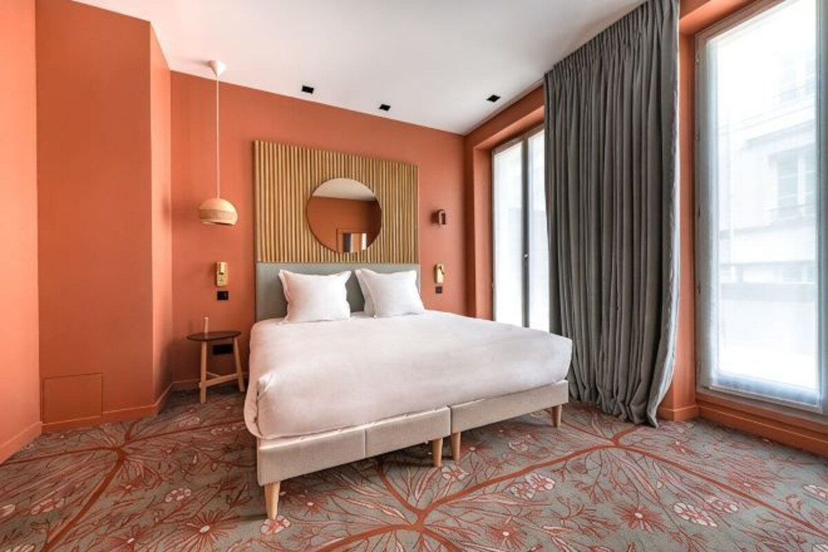 A hotel room with a large bed, a small bedside table, a mirror, and a view of the city through a window.