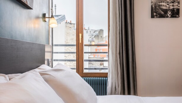Una habitación de hotel con una cama, un ventanal con vistas a edificios, una lámpara de pared y un radiador.