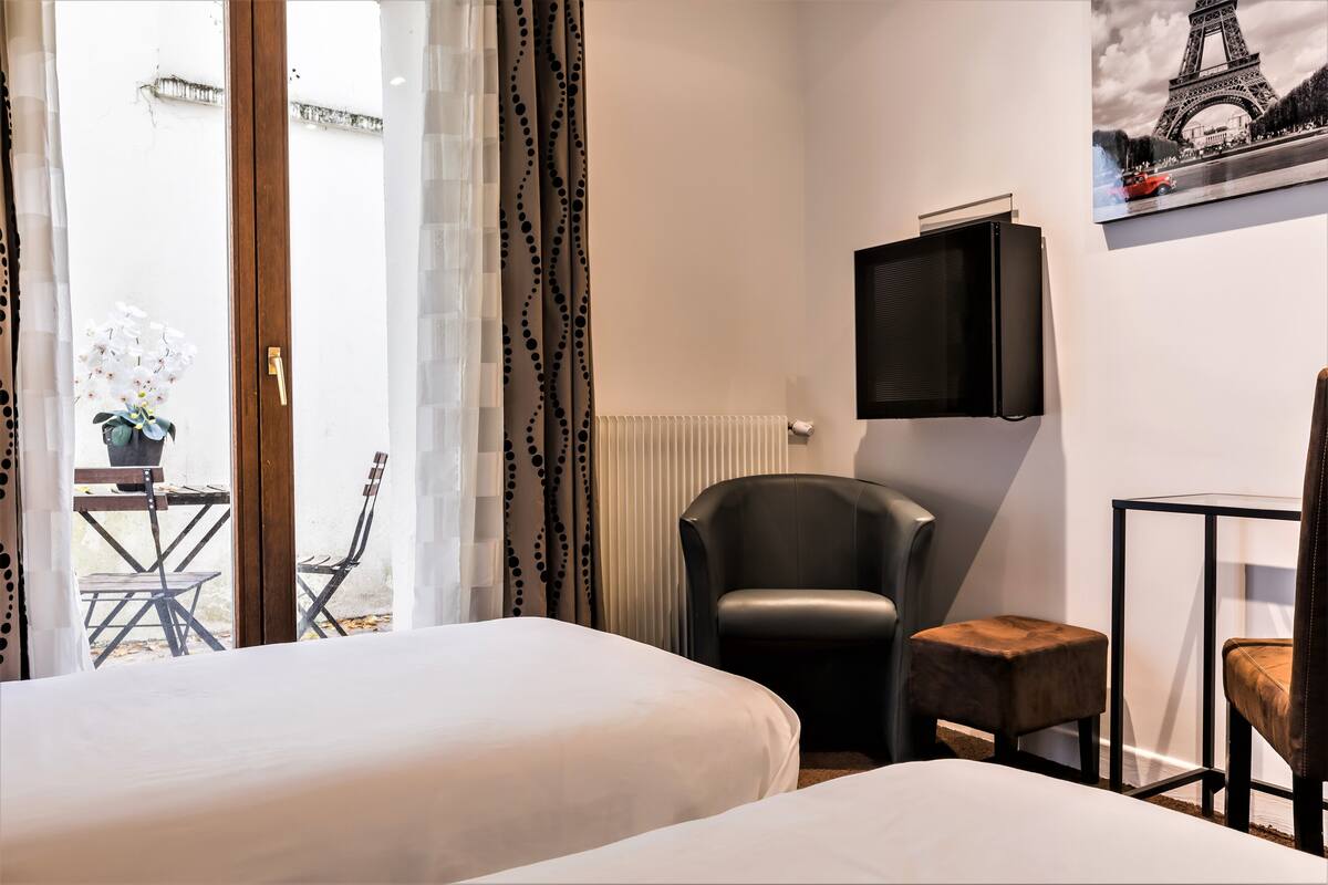 A hotel room with two beds, a black leather chair, a small stool, a television, and a view of the Eiffel Tower through a window.