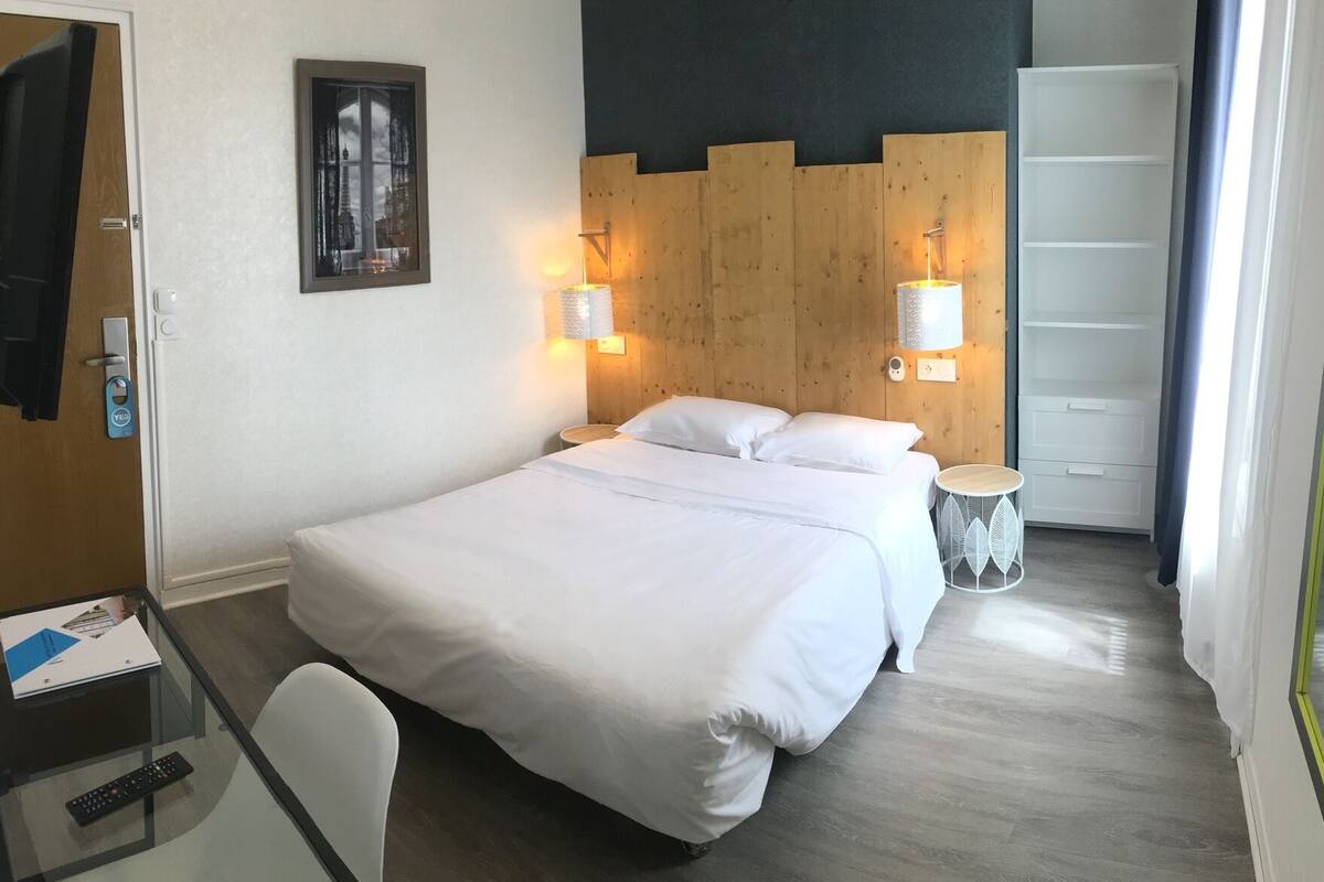 A hotel room with a large bed, two bedside lamps, a small round table, a chair, a desk with a computer and a remote control, and a wardrobe.
