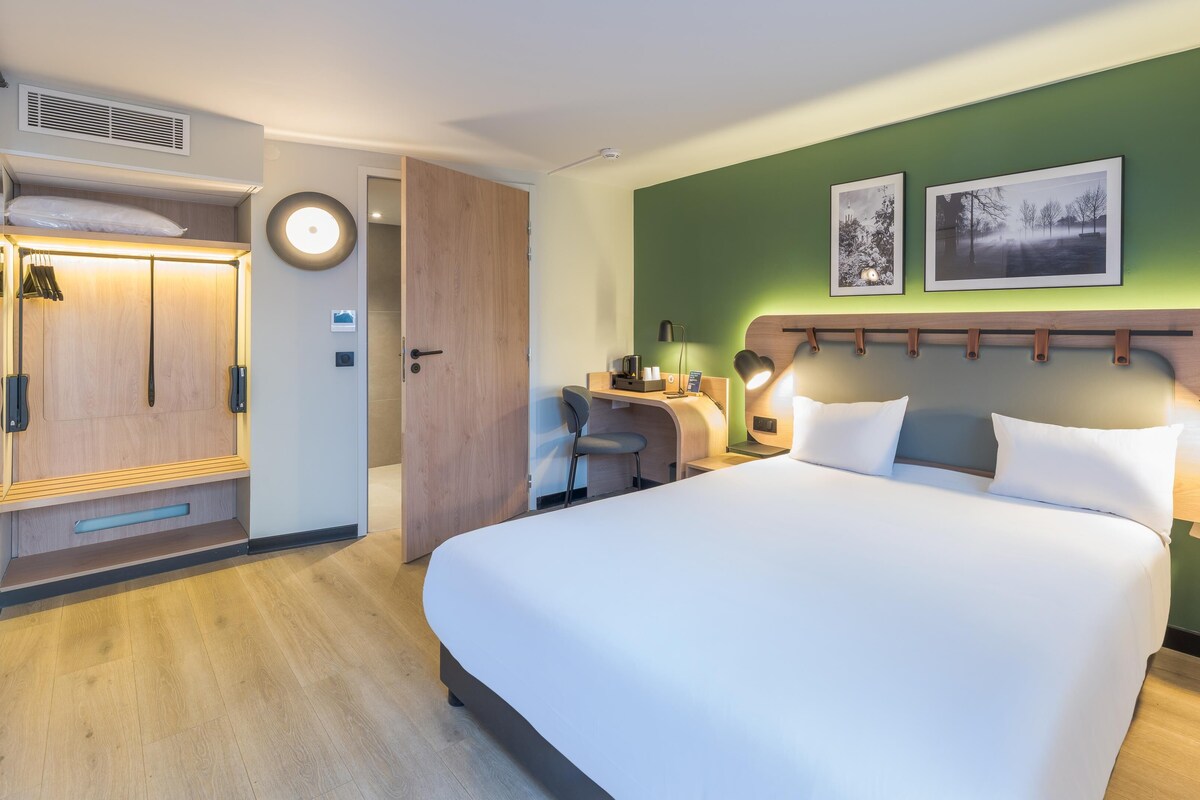 Habitación de hotel con cama, escritorio, silla, armario y cabecera con lámpara.