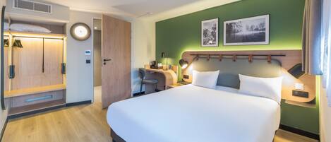 Habitación de hotel con cama, escritorio, silla, armario y cabecera con lámpara.