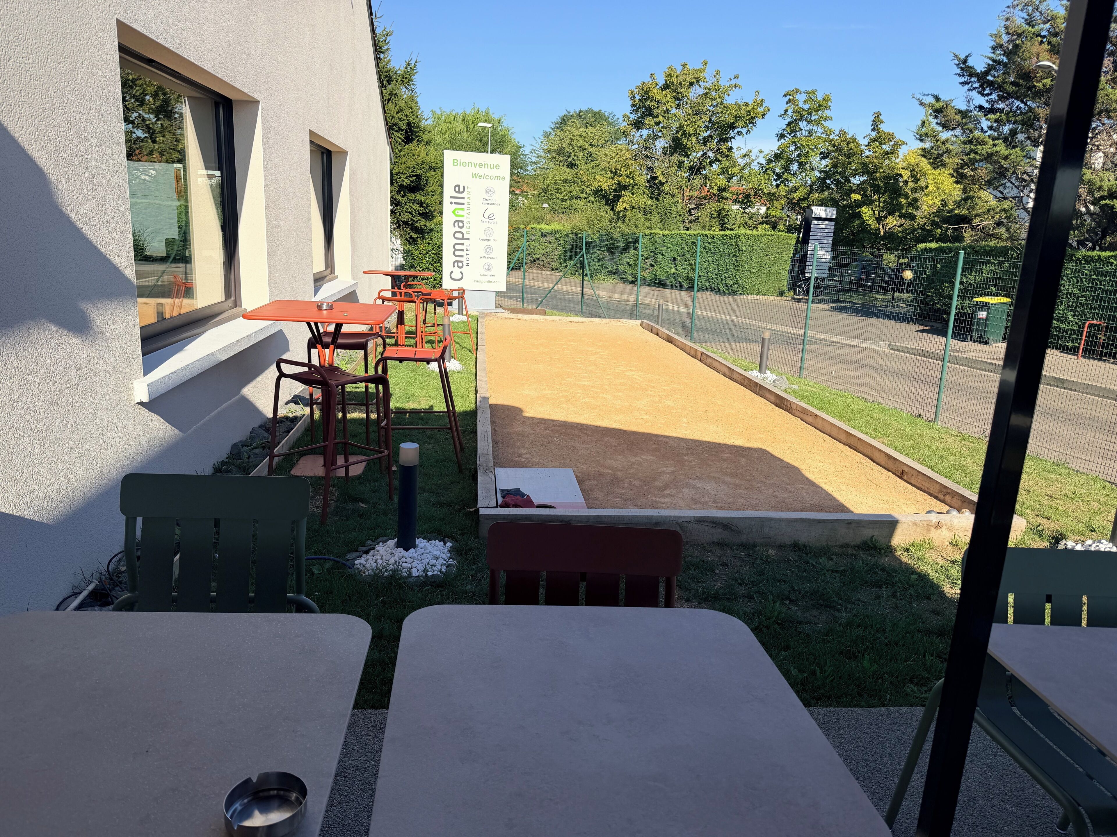 An outdoor seating area with tables and chairs, a sign indicating a welcome message, and a fenced garden.