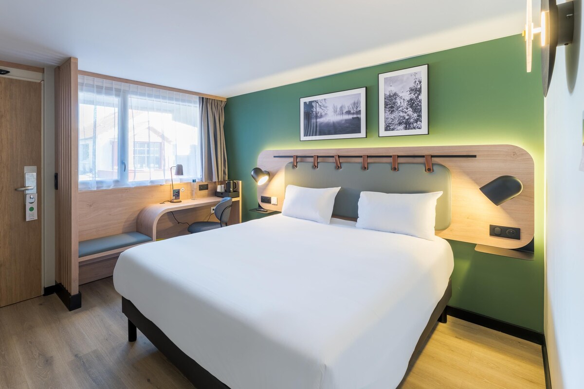 Habitación de hotel con cama, escritorio, silla, ventana con cortinas y dos cuadros en la pared.