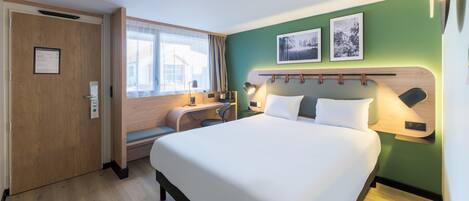 A hotel room with a bed, a desk, a chair, a window with curtains, and two framed pictures on the wall.