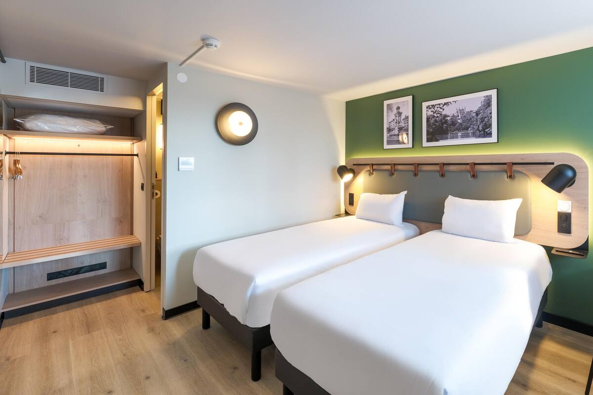 Een hotelkamer met twee bedden, een kledingkast, een bureau met stoel, en twee ingelijste foto's aan de muur.