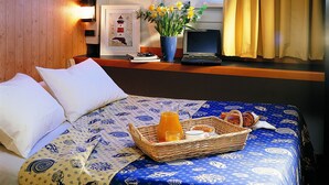 Un lit bien fait, recouvert d’une couvre-lit aux motifs bleus et jaunes, un panier en osier contenant des articles pour le petit-déjeuner, et un ordinateur portable posé sur un bureau agrémenté d’une photo encadrée et de fleurs.