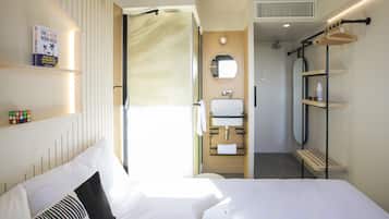 A hotel room with a bed, a bathroom with a mirror, and a shelf with a book and a water bottle.