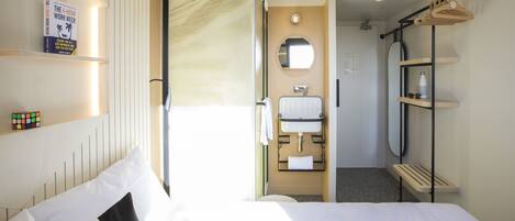 Una habitación de hotel con cama, baño con espejo y estante con un libro y una botella de agua.