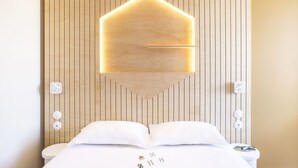 Une chambre d’hôtel avec un mur lambrissé en bois, un lit avec une literie blanche et un luminaire géométrique original placé au-dessus du lit.
