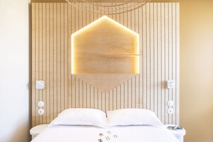 Une chambre d’hôtel avec un mur lambrissé en bois, un lit avec une literie blanche et un luminaire géométrique original placé au-dessus du lit.