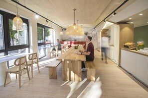 Un restaurant moderne avec des tables et des chaises en bois, un bar proposant des fruits et des boissons, et de grandes fenêtres laissant entrer la lumière naturelle.