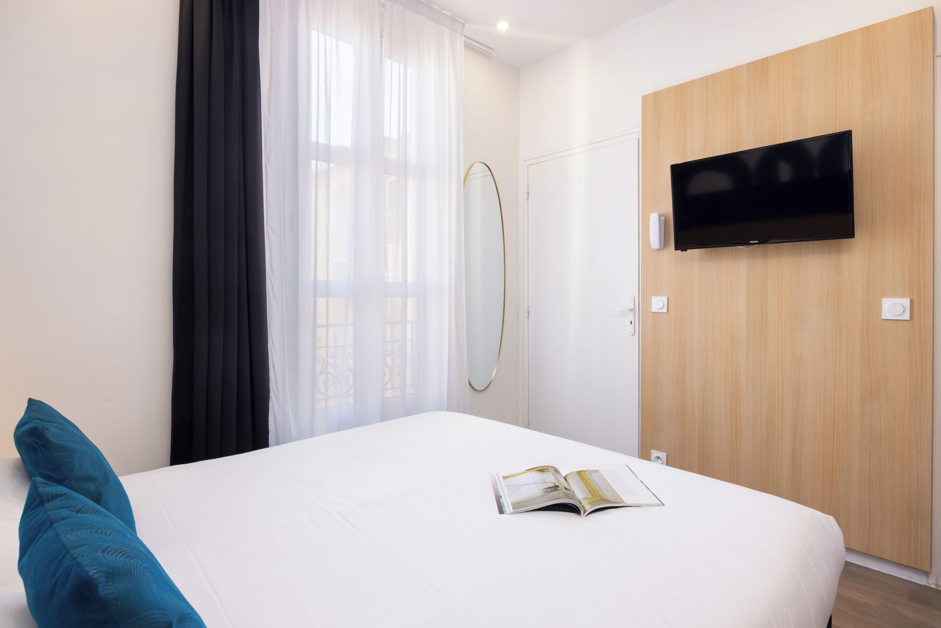 A hotel room with a bed, a TV mounted on the wall, a window with sheer curtains, and a wooden door.