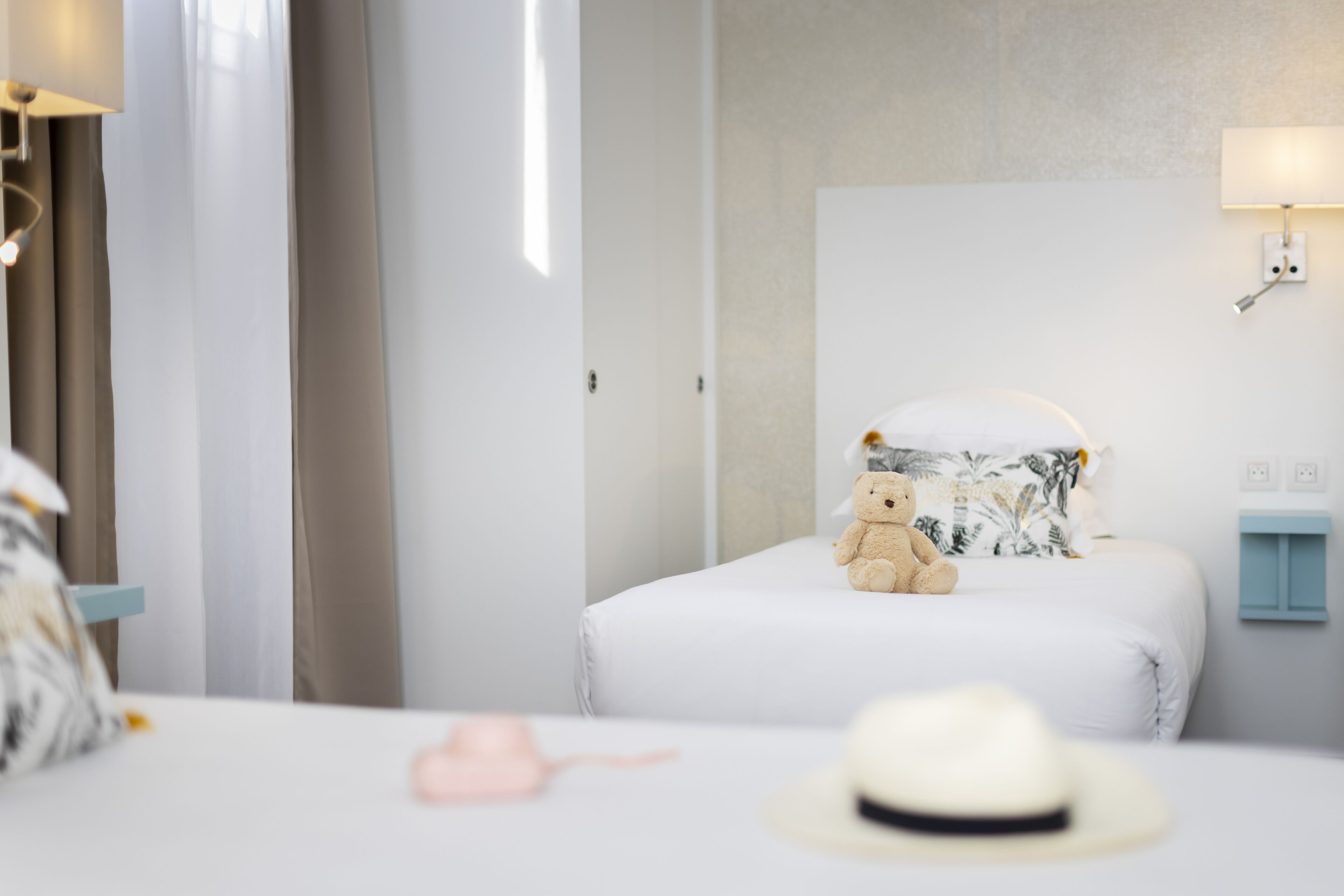 A neatly made bed with a white comforter, a teddy bear, and a white hat on it.