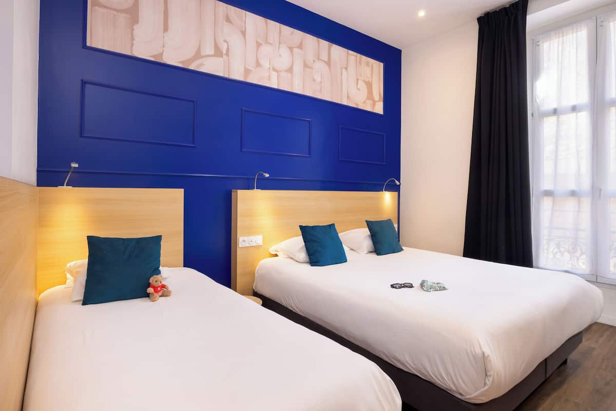Deux lits avec des têtes de lit, un mur orné d’un motif géométrique et une fenêtre avec des rideaux.