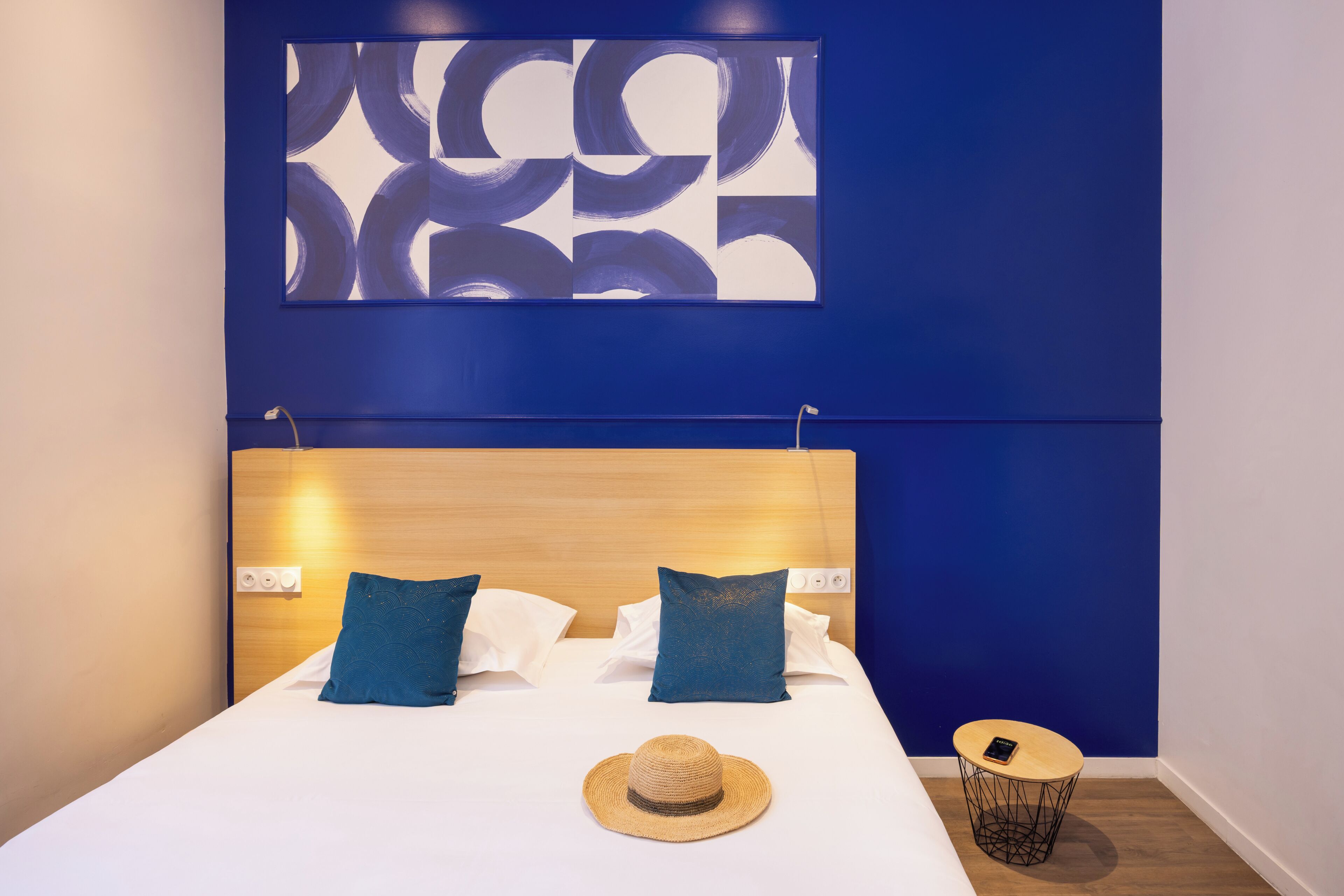 A neatly made bed with two pillows, a hat on the bed, and a small round table with a phone on it.