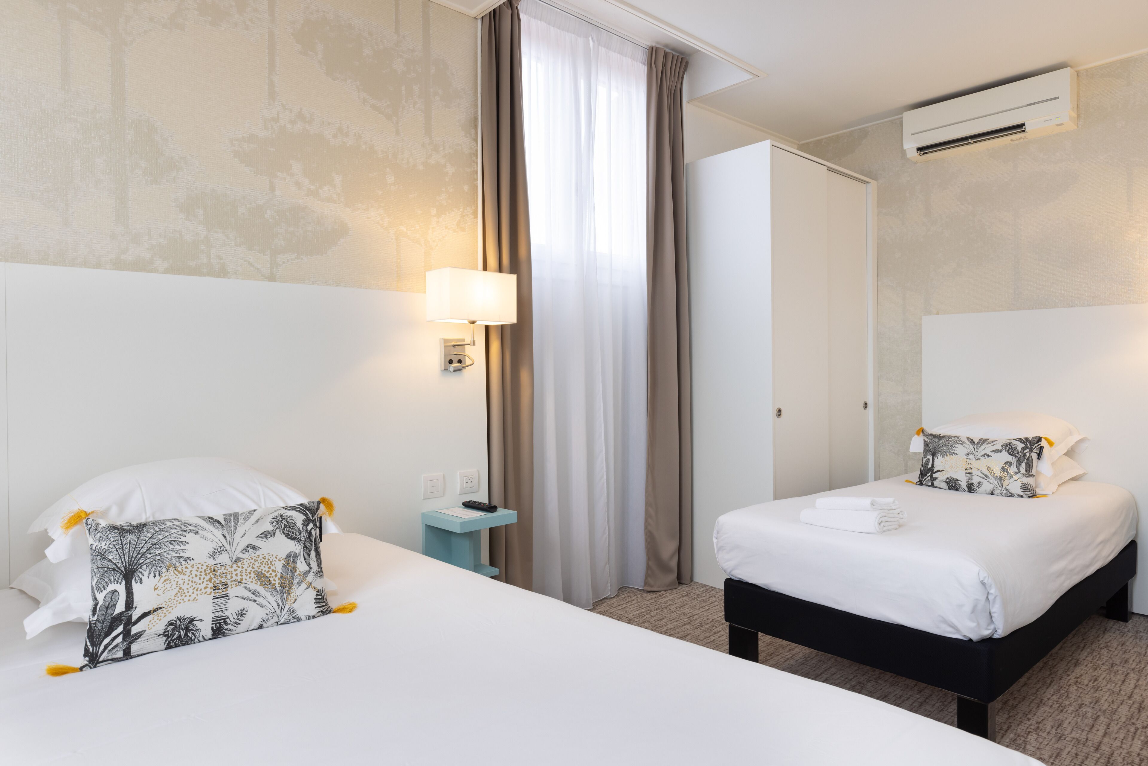 A hotel room with two beds, a bedside table with a lamp, a window with curtains, and an air conditioning unit.