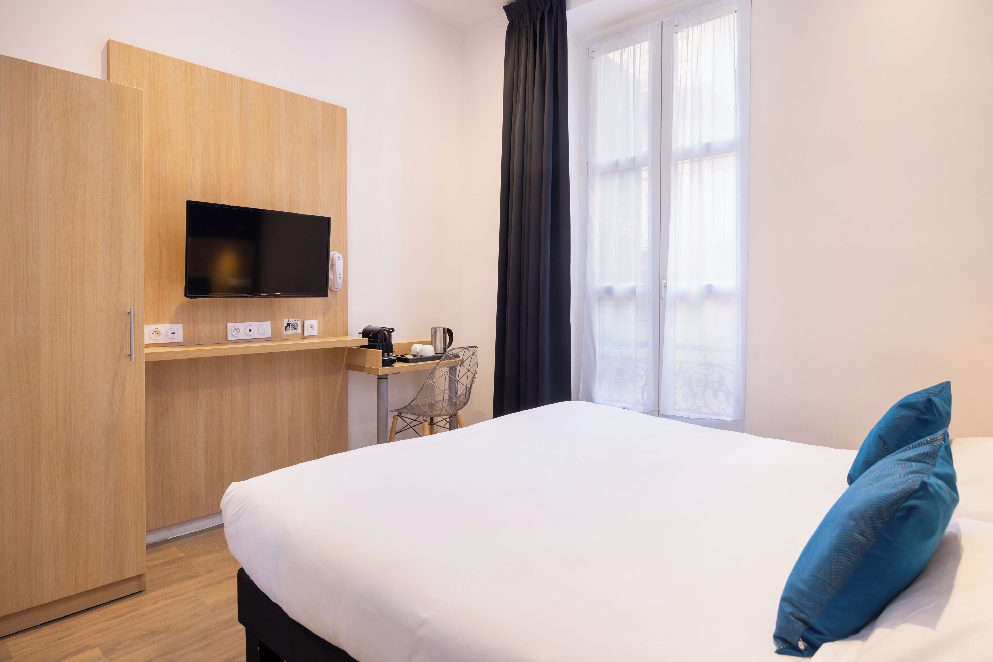 A hotel room with a bed, a TV mounted on a wooden panel, a desk with a chair, and a window with curtains.