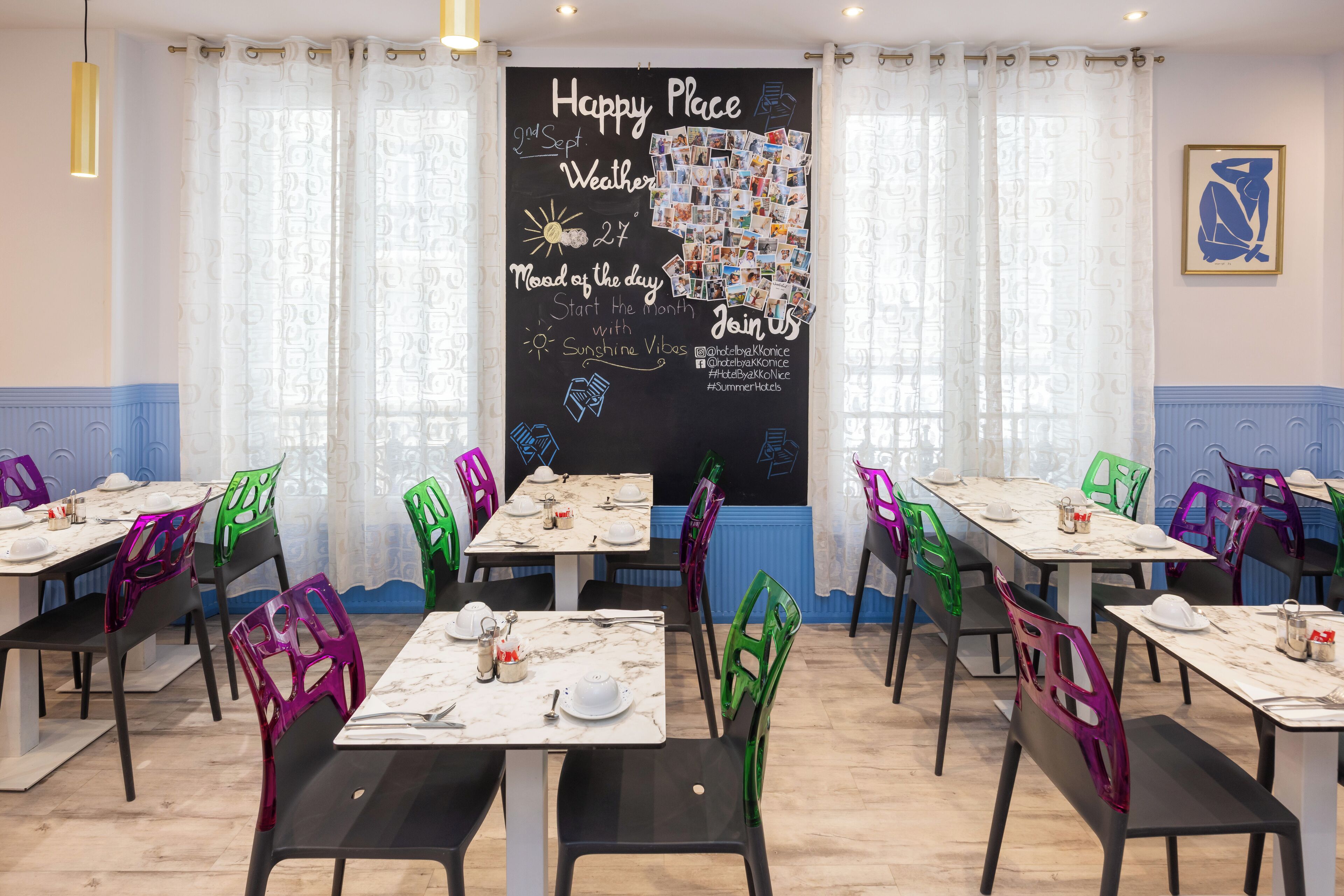 Un espace repas avec des tables disposées pour accueillir quatre personnes, des chaises dotées de dossiers ajourés et un tableau noir orné de photos et de textes.