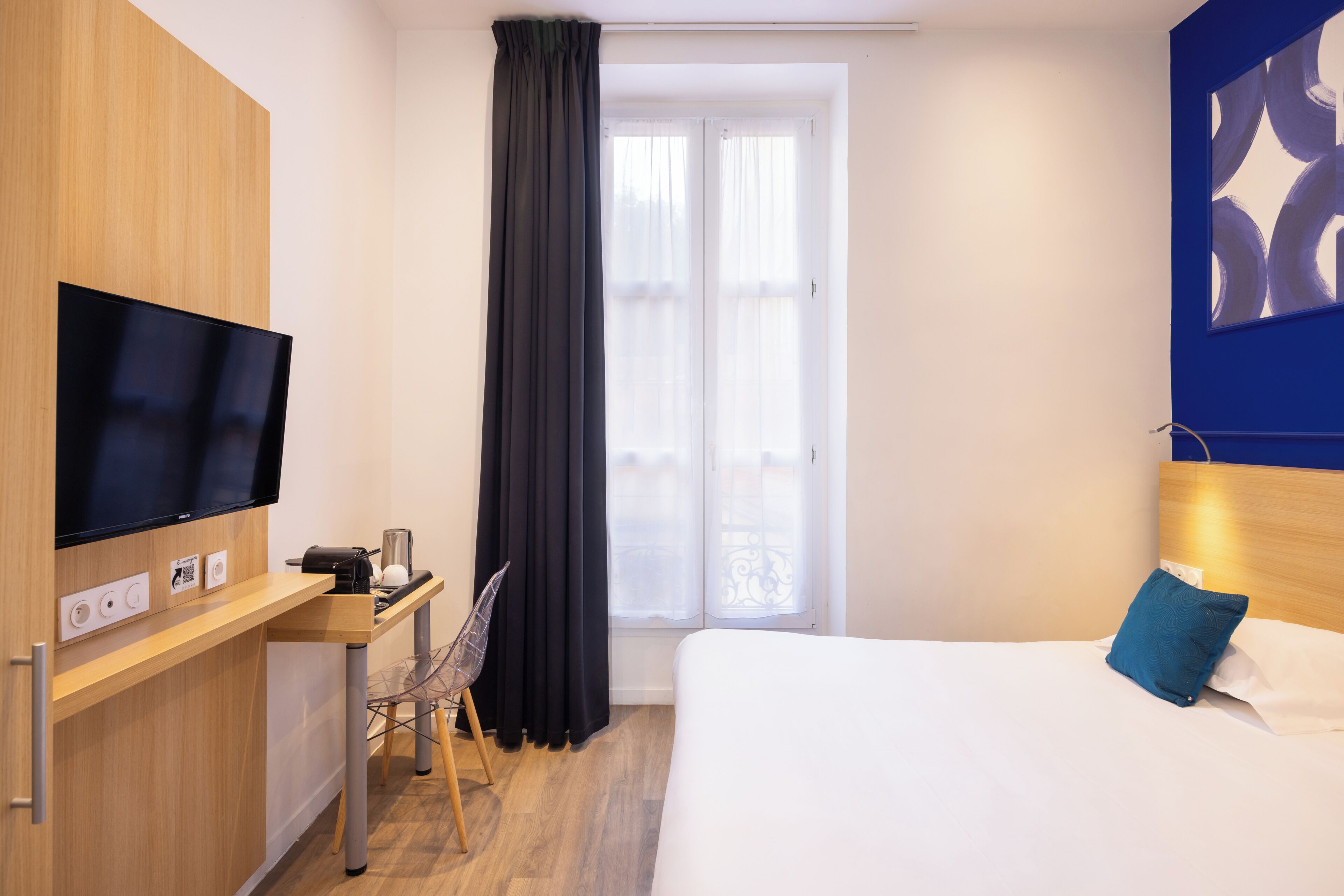 A hotel room with a bed, a desk with a chair, a television, and a window with curtains.