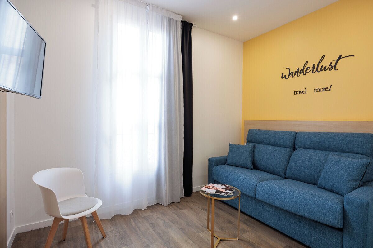 Ein modernes Wohnzimmer mit einem blauen Sofa, einem weißen Sessel, einem kleinen runden Tisch mit einer Zeitschrift und einer Wand mit dem Schriftzug 'Wanderlust travel more!'