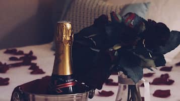 Ein Bett mit weißer Bettdecke und verstreuten Rosenblättern. Auf dem Bett steht eine Champagnerflasche in einem Metallkübel. Ein Rosenstrauß aus dunkelgrünen Rosen befindet sich in einer Vase auf dem Bett.
