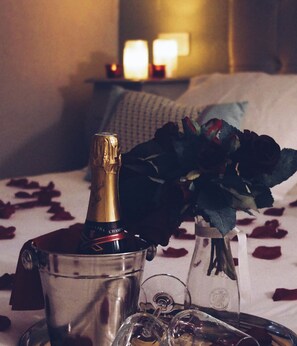 A bed with a white bedspread and rose petals scattered on it. A champagne bottle is placed in a metal bucket on the bed. There is a bouquet of dark green roses in a vase on the bed.
