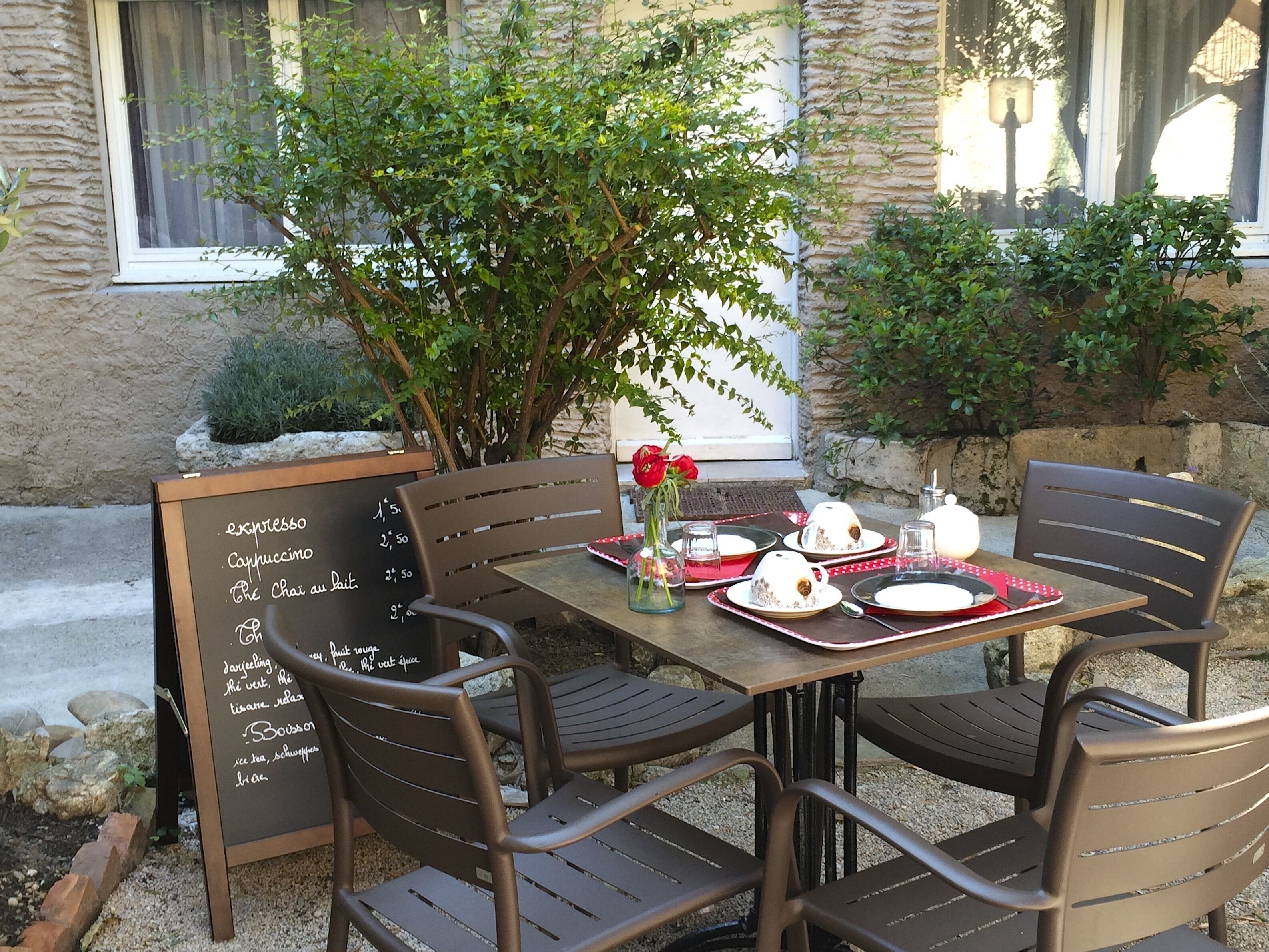 Un espace de restauration en plein air avec une table en bois et quatre chaises, un tableau noir servant de menu et un vase rempli de fleurs rouges.