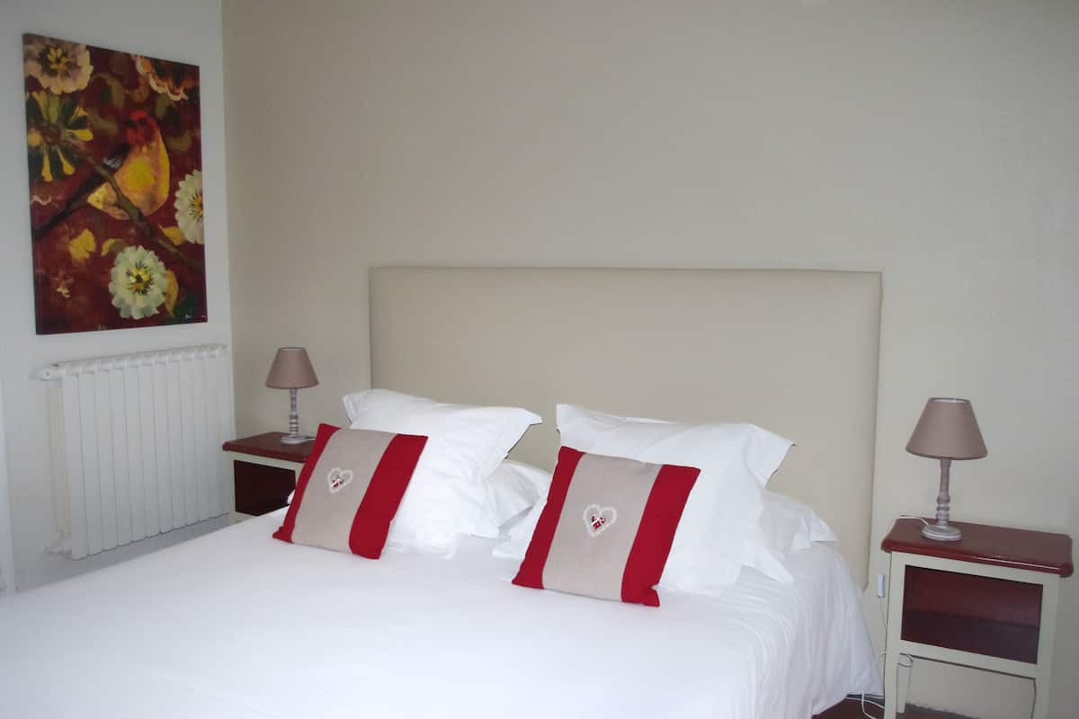 Una cama bien hecha con sábanas blancas y dos almohadas, cada una con un diseño rojo y beige. Hay dos mesitas de noche con lámparas, y un cuadro con motivos florales en la pared.