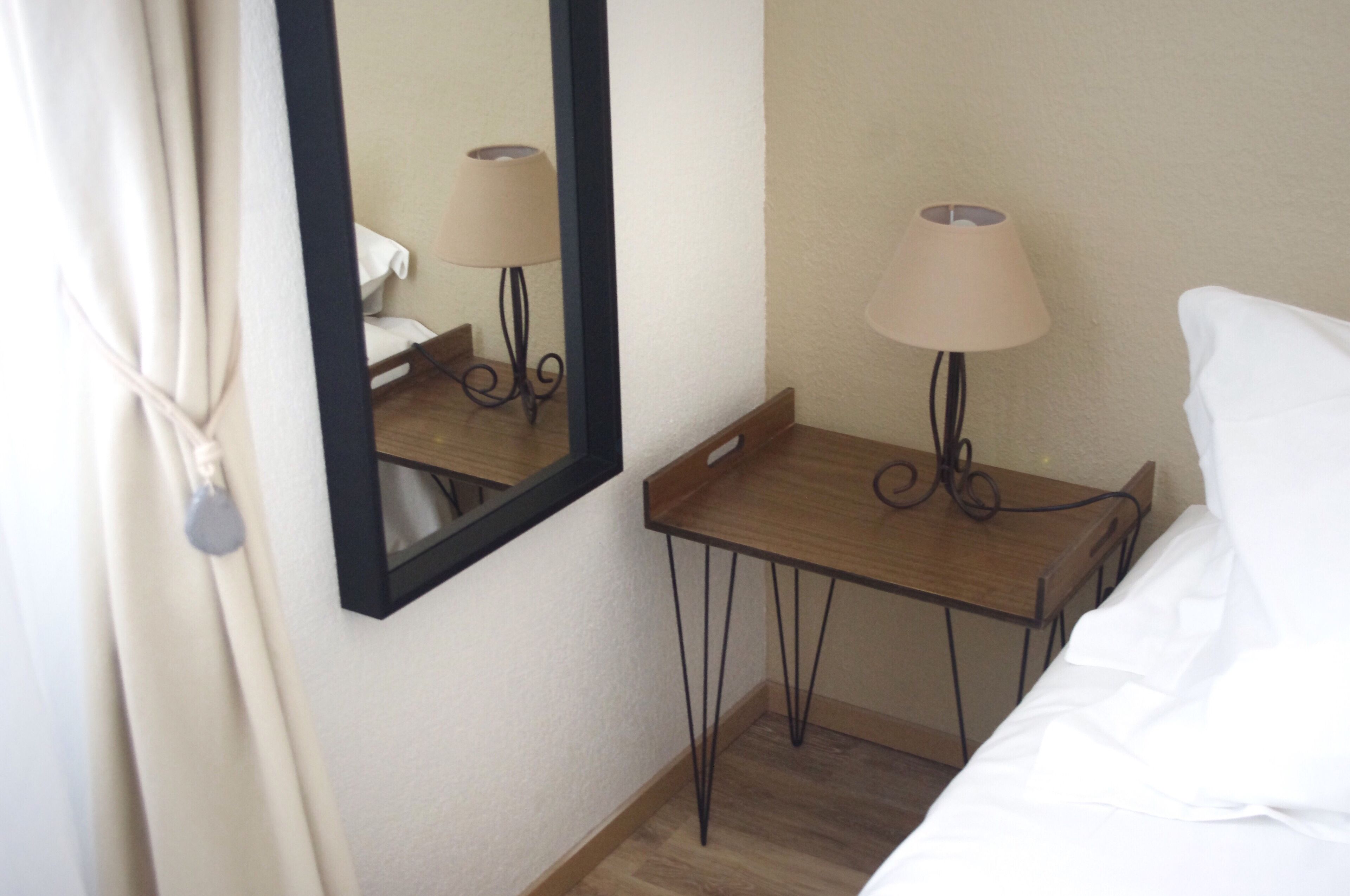 Habitación de hotel con cama, mesita de noche con lámpara, espejo y ventana con cortinas.
