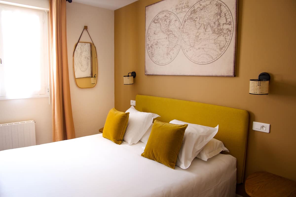 Una cama bien tendida con almohadas blancas y azafrán, un cabecero amarillo y una obra de arte con un mapa del mundo en la pared.