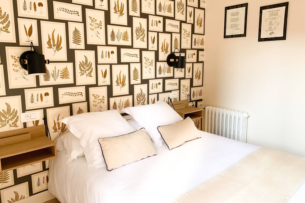Un dormitorio con cama, almohadas blancas, mesita de noche, lámpara de pared y láminas botánicas enmarcadas.