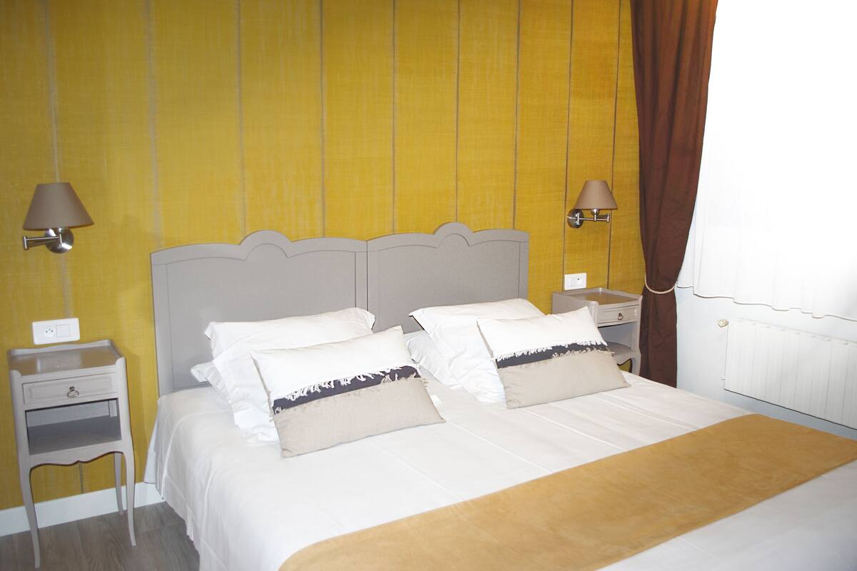 Una cama bien hecha con ropa blanca y dos almohadas, una mesita de noche con una lámpara, y una pared con un panel amarillo.