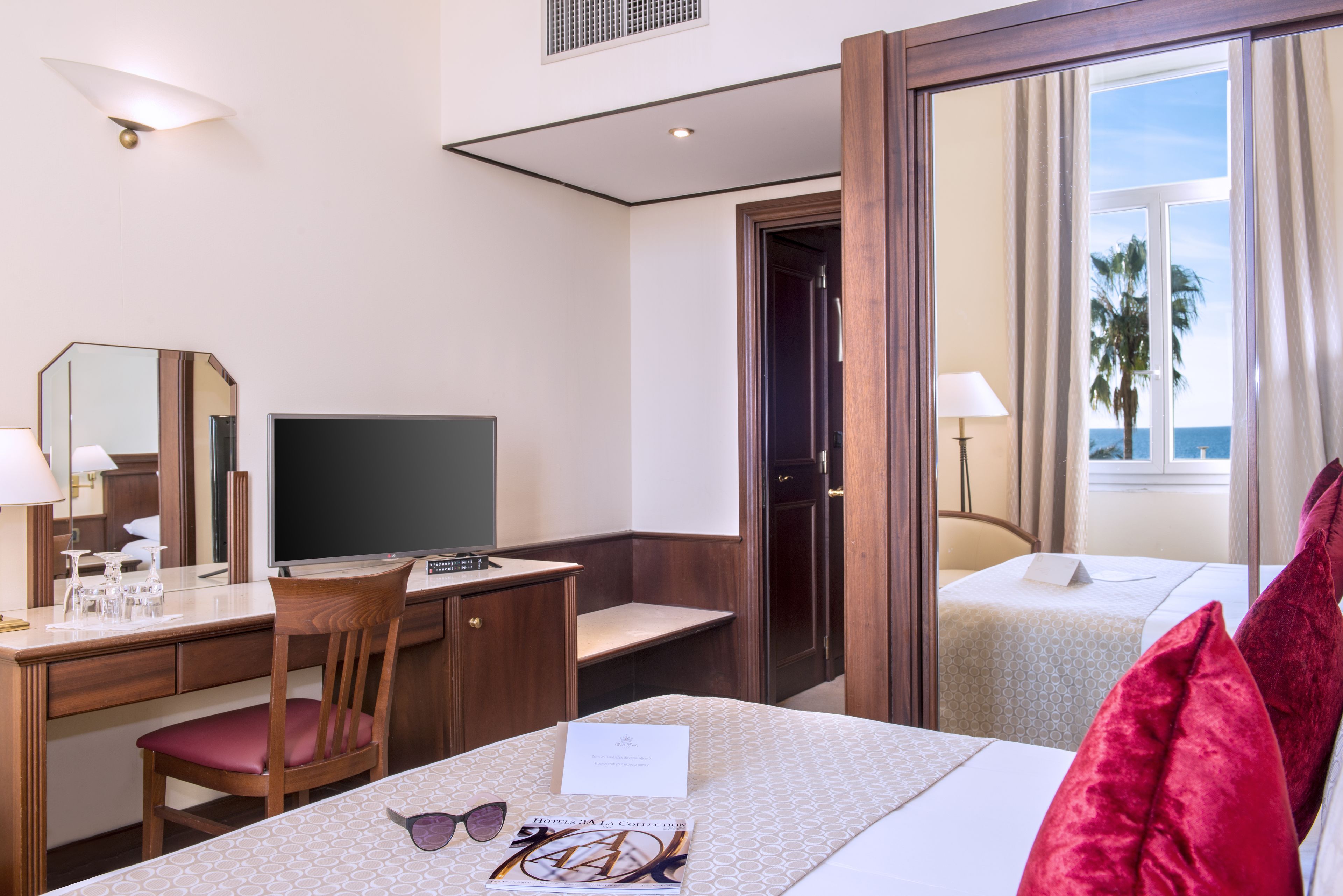 Habitación de hotel con escritorio, cama, televisión, ventana con vista al mar y balcón.