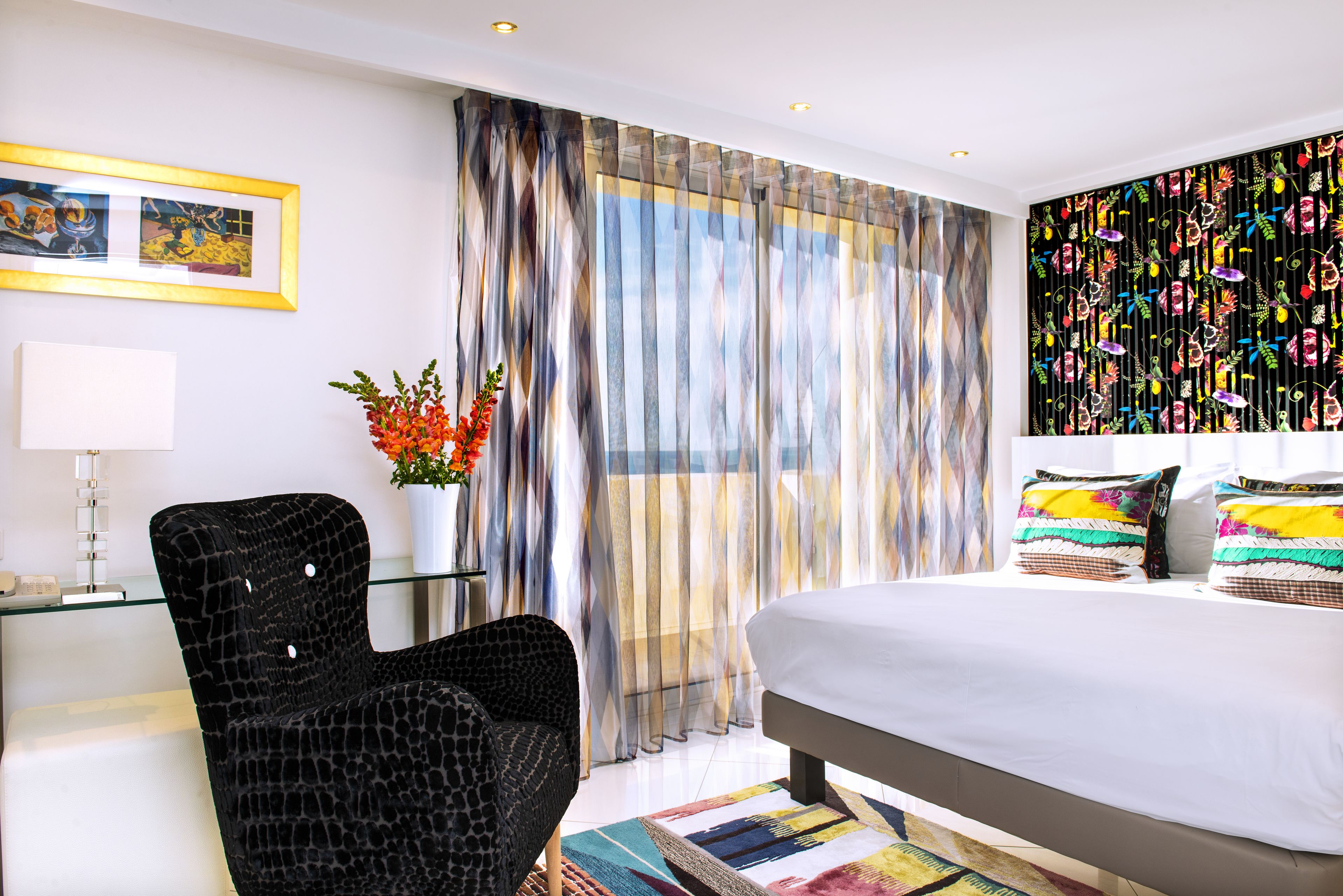 Une chambre d’hôtel moderne avec un grand lit, un bureau en verre, un fauteuil noir et un mur décoré d’une œuvre abstraite aux couleurs vives.