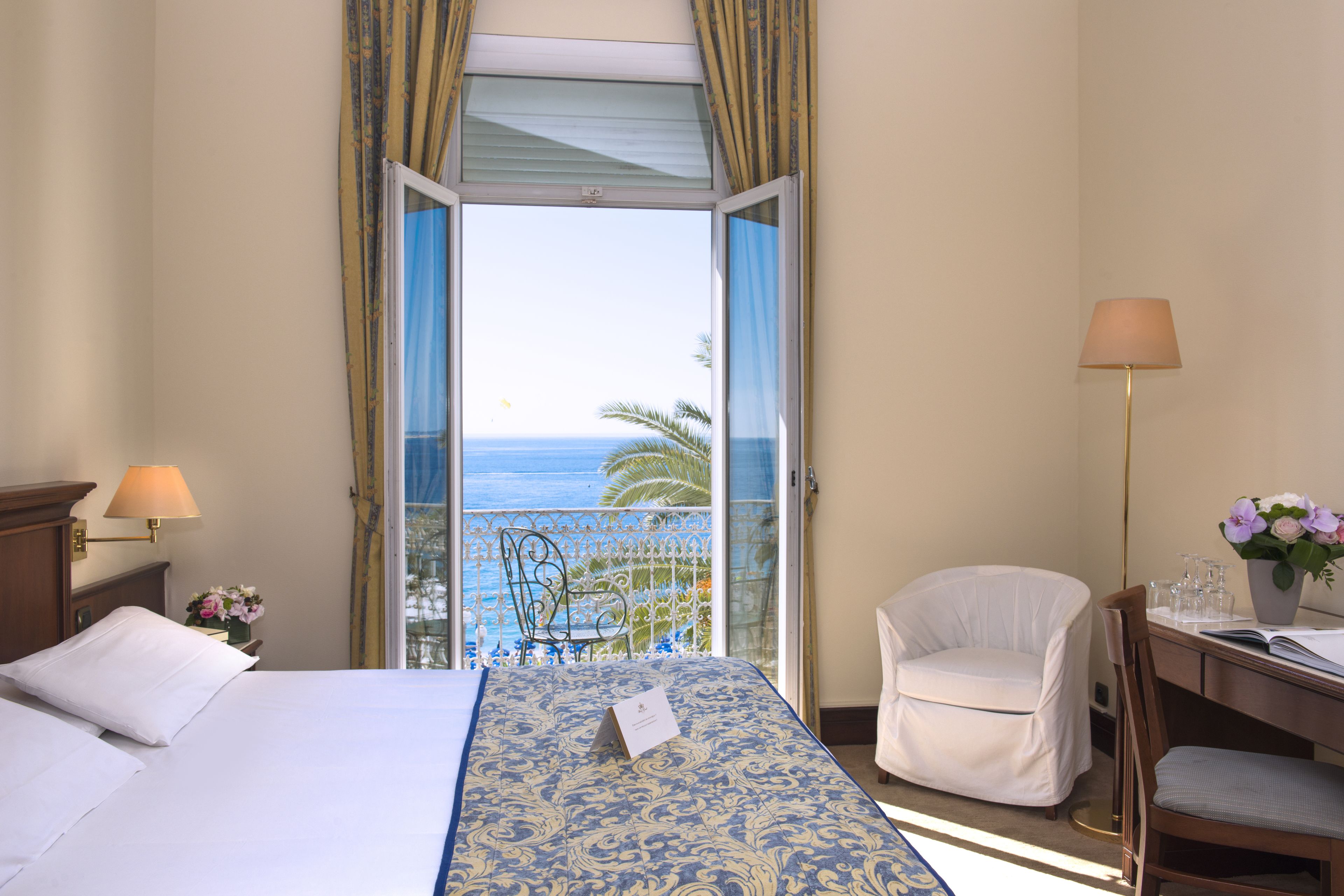 Una habitación de hotel con una cama, un escritorio con una silla, una lámpara y vista al océano.