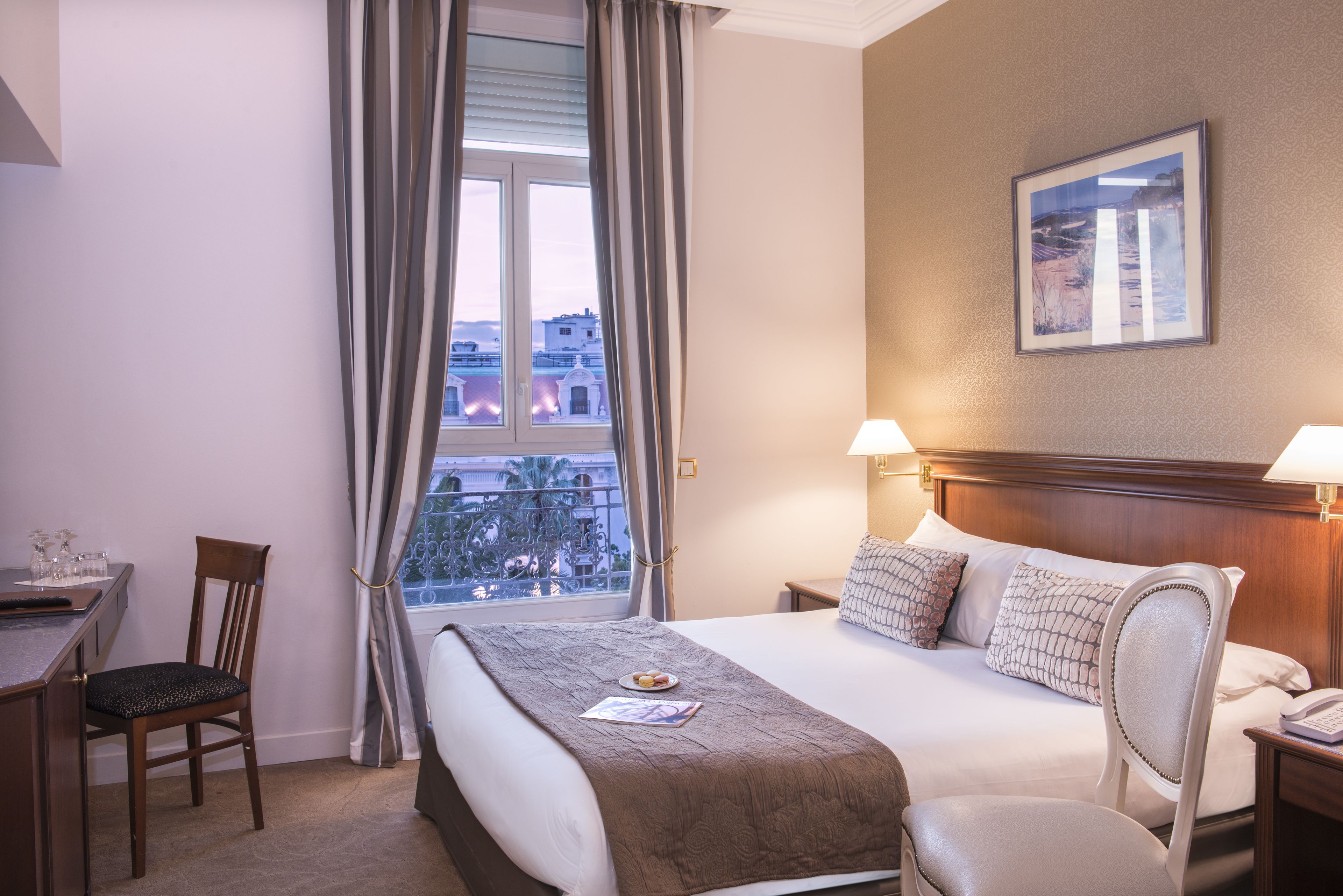A hotel room with a bed, a desk, a chair, a window with curtains, and a painting on the wall.