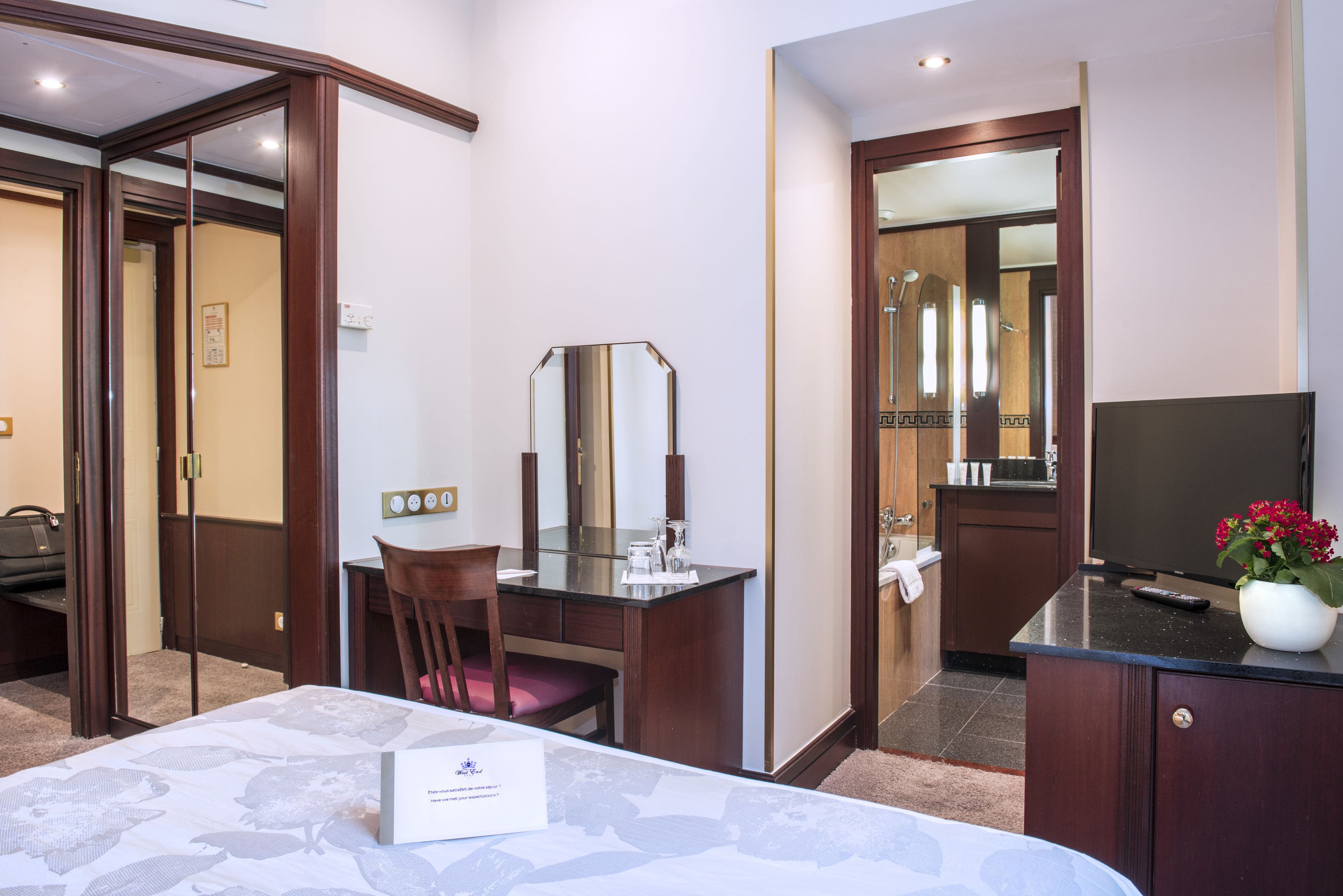 A hotel room with a bed, a desk with a chair, a mirror, a TV, and a bathroom visible through a door.