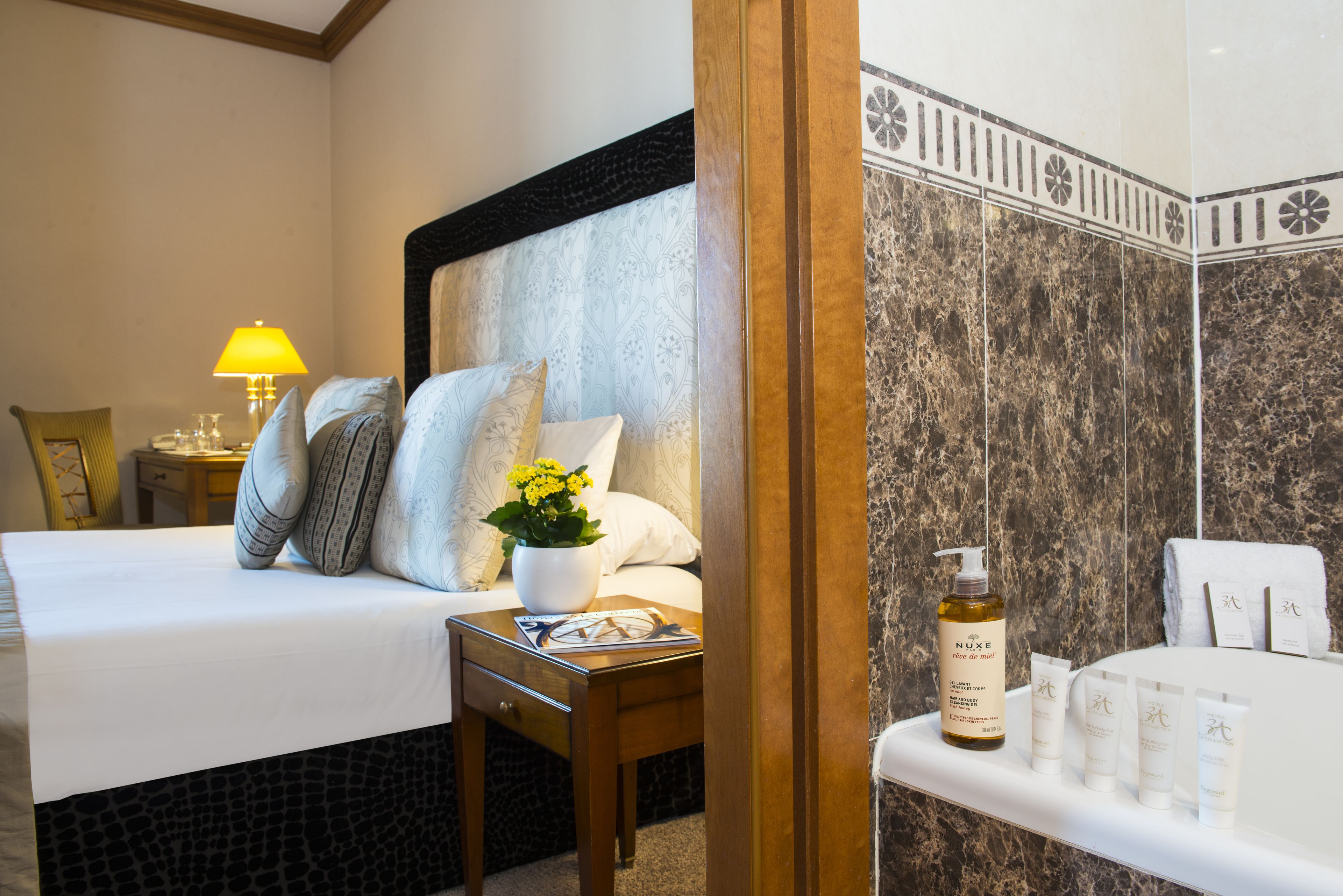A hotel room with a bed, bedside table with a lamp, a nightstand, a bathroom with a bathtub, and toiletries on the sink.