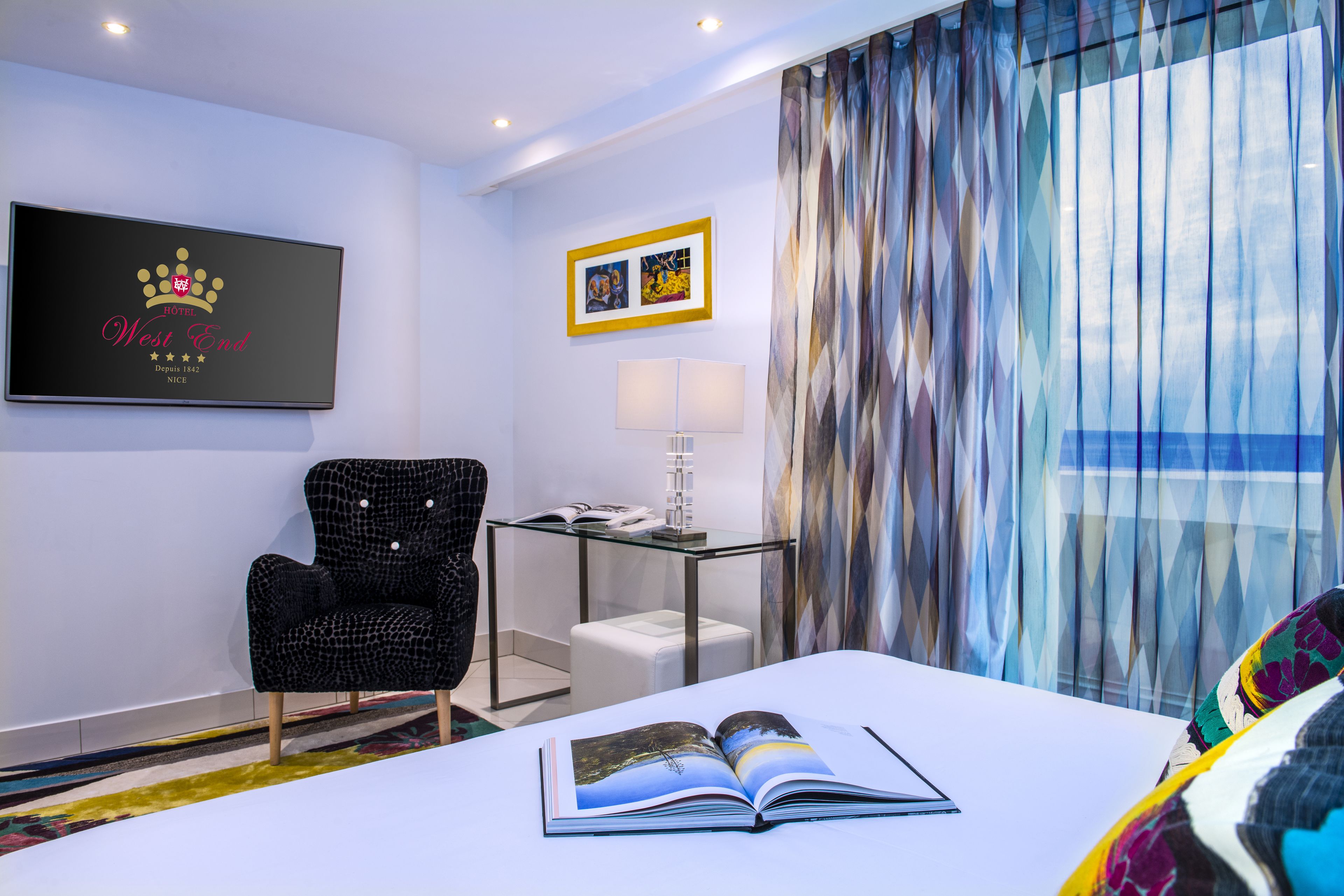Habitación de hotel con cama, una silla, una mesita con lámpara, un cuadro en la pared y una ventana con cortinas.
