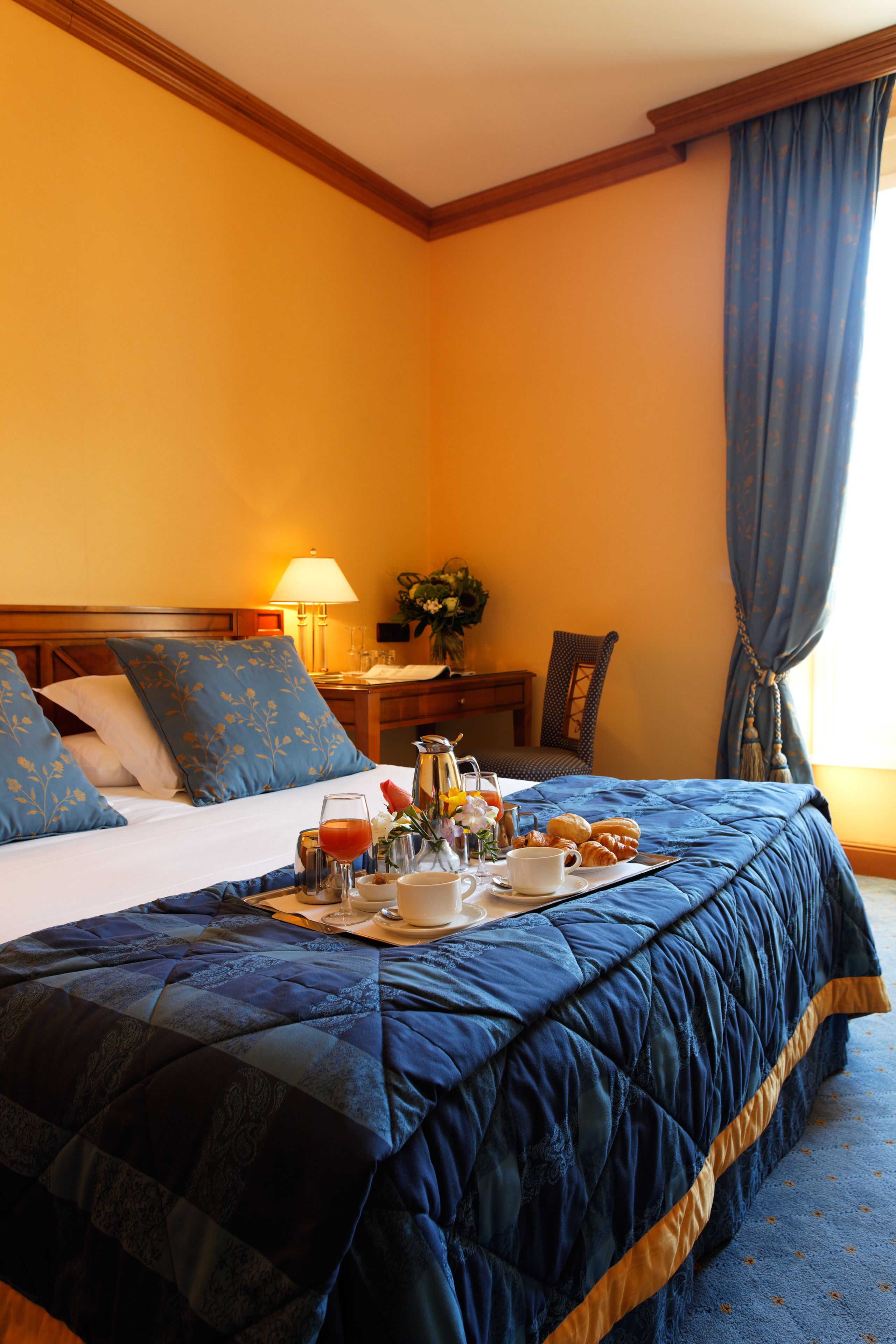 Een hotelkamer met een groot bed, een nachtkastje met een lamp, een stoel en een dienblad met ontbijtartikelen.