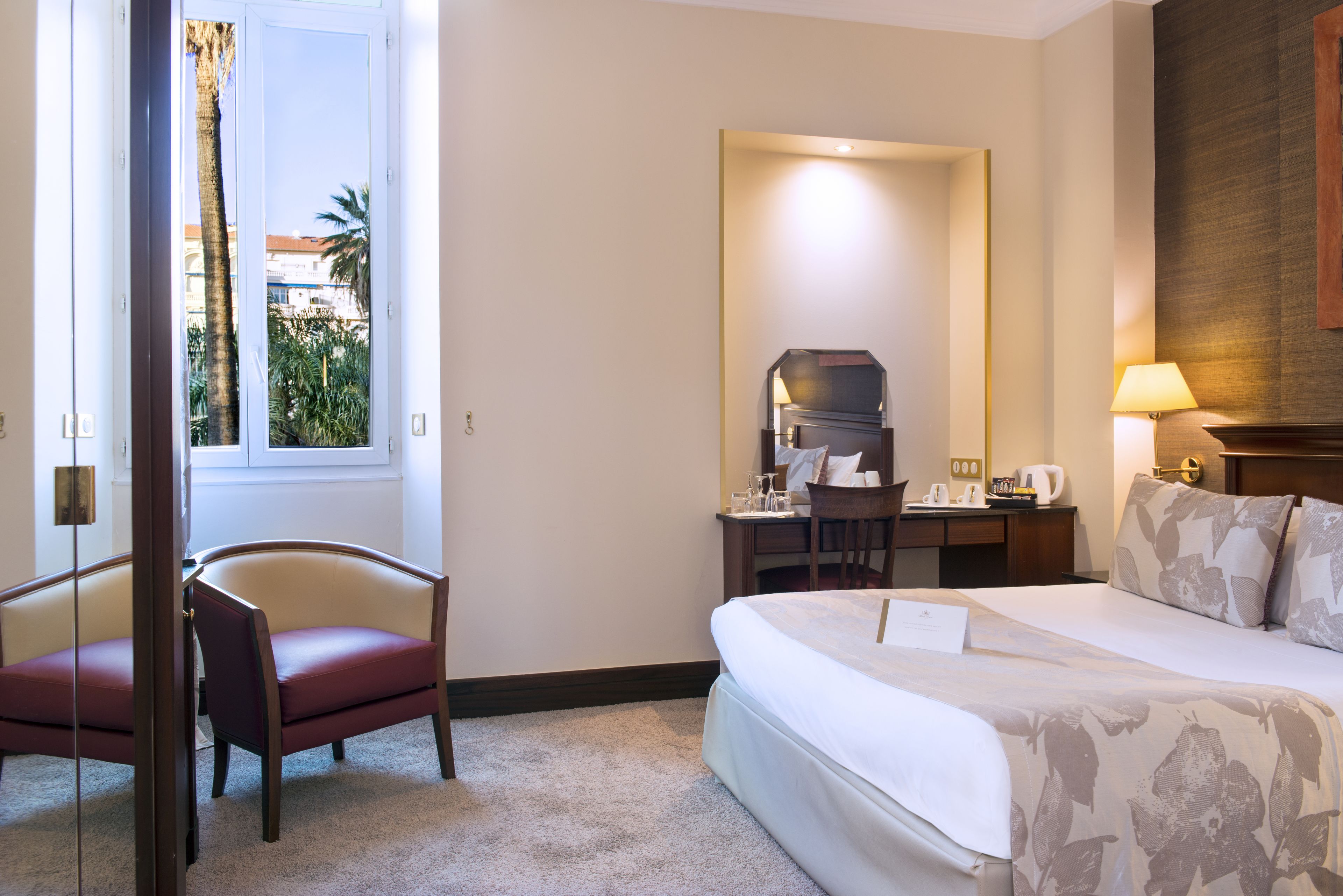 Habitación de hotel con cama, escritorio con silla, ventana con vista a palmeras y una lámpara.