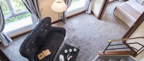 A hotel room with a black patterned sofa, a small coffee table with cups and a book, a standing lamp, and a view of the outdoors through large windows.