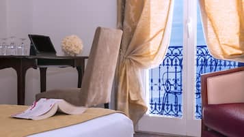 A hotel room with a bed, a desk with a laptop, a chair, and a view of a balcony with blue railing.