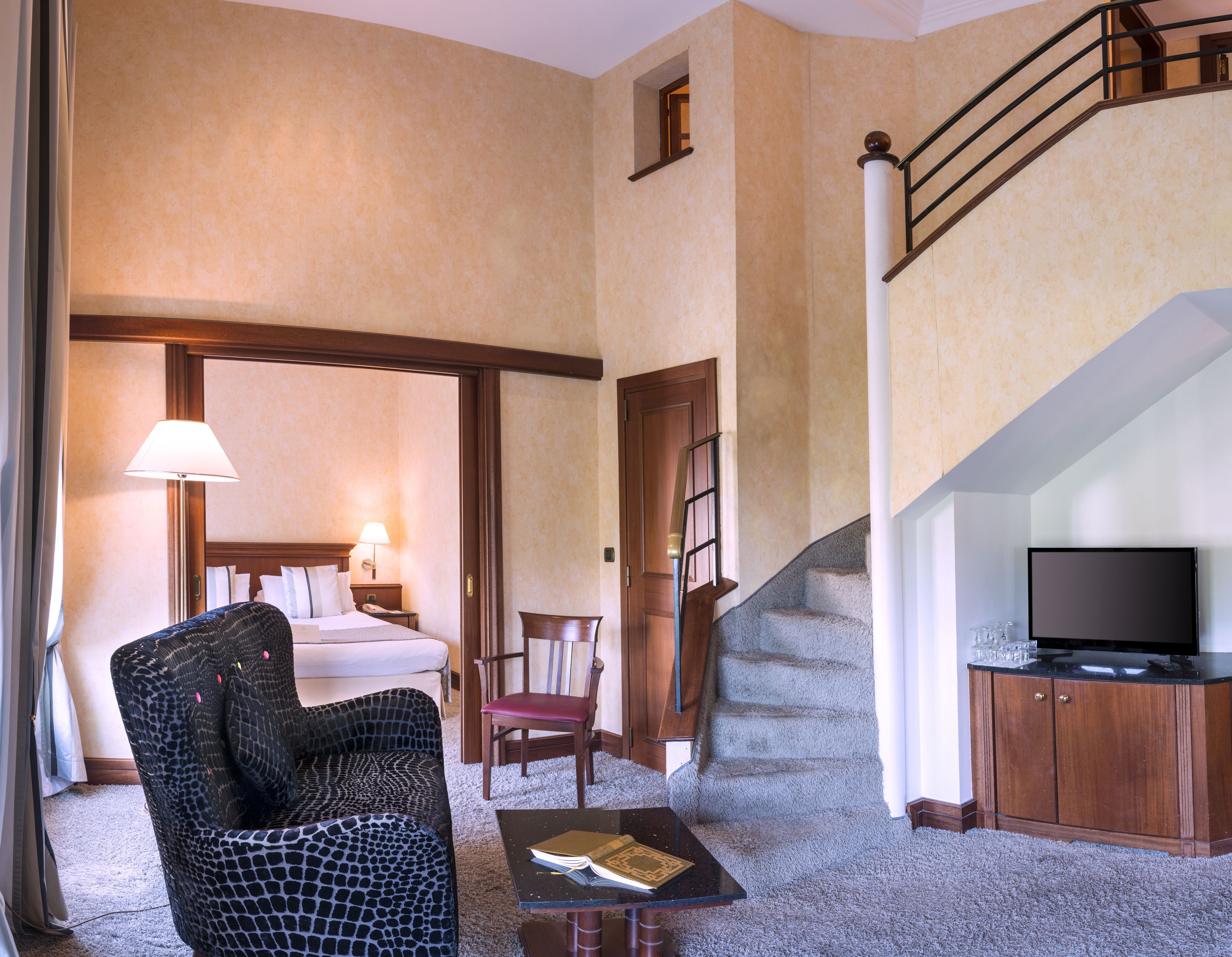 Habitación de hotel con cama, una silla, una mesa con un libro, televisor y escalera.